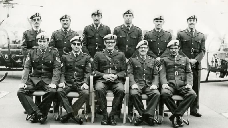 Black and white photo of eleven men in military uniforms outside with helicopters in background behind them. Five sit at front and six stand behind in rows. 