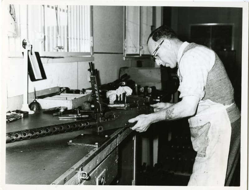 Black and white photo of a man in an apron fixing a large gun at a workshop station. Other pieces of metal and materials on the station. 